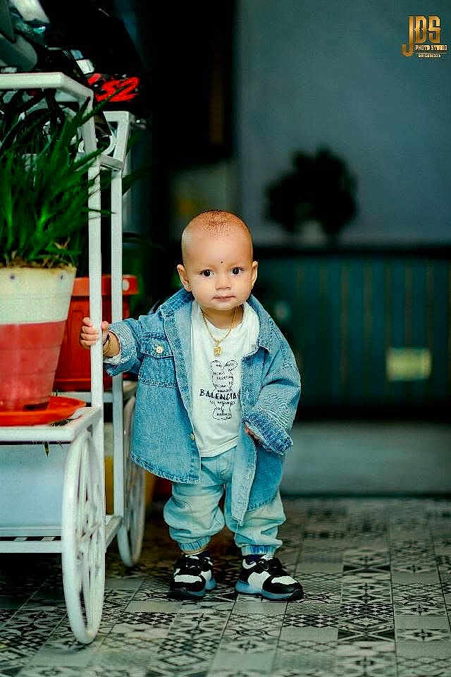 Jarvis is registered to the contest to win money with this photo: toddler, child, denim_jacket, sneakers, indoor, potted_plant, cart, floor_tiles, curious, standing, casual_clothing, portrait, young_child, cute, fashionable, jewelry, baby, person, floor, background_blur
