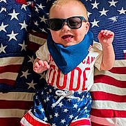 Ryker is registered to the contest to win money with this photo: american_flag, baby, baby_clothing, bib, blanket, blue, child, fashion, happy, indoor, infant, lying_down, patriotic, portrait, red, smiling, stars, stripes, sunglasses, white