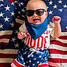 american_flag, baby, baby_clothing, bib, blanket, blue, child, fashion, happy, indoor, infant, lying_down, patriotic, portrait, red, smiling, stars, stripes, sunglasses, white