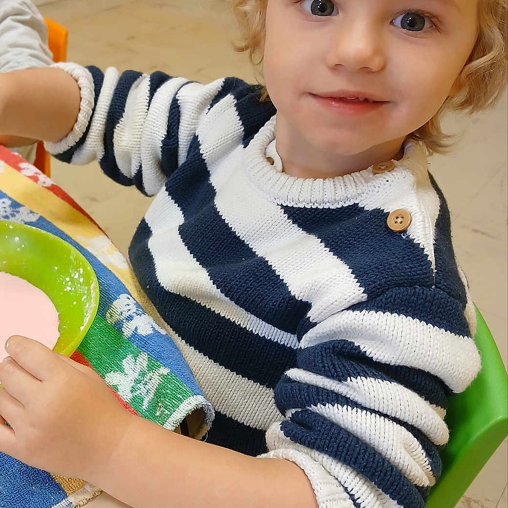 Alissio a rejoint le concours — aidez-le/la à gagner de superbes lots ! blonde_hair, blue_eyes, bowl, child, cozy, curly_hair, cute, green_chair, indoor, person, pink_liquid, playful, portrait, sitting, smiling, striped_sweater, table, towel, yellow_wall, young