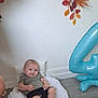 baby, infant, blanket, balloon, number, blue, floor, wooden_floor, wall, decor, autumn_leaves, pillow, clothing, shorts, tshirt, smile, face, person, indoors, celebration