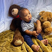 Isaiah is registered to the contest to win money with this photo: toddler, child, teddy_bear, plush_toy, blanket, striped_shirt, blue_pants, barefoot, indoor, portrait, curious_expression, soft_lighting, white_drapery, brown_teddy_bear, golden_blanket, sitting, cute, young_child, cozy, background