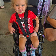 Emryn is registered to the contest to win money with this photo: child, toddler, stroller, smiling, portrait, outdoor, red_shirt, blue_shoes, backpack, harness, seated, blonde_hair, blue_eyes, playground, concrete, parent, casual_clothing, wheelchair_style_stroller, happy, photography