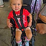 child, toddler, stroller, smiling, portrait, outdoor, red_shirt, blue_shoes, backpack, harness, seated, blonde_hair, blue_eyes, playground, concrete, parent, casual_clothing, wheelchair_style_stroller, happy, photography