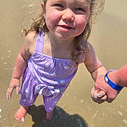 Emryn is registered to the contest to win money with this photo: adult_hand, beach, blue_bracelet, child, closeup, fish_pattern, girl, holding_hand, outdoor, portrait, purple_outfit, seawater, shadow, shoreline, smiling, summer, sunlight, toddler, wet, windblown_hair