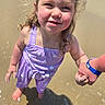child, girl, toddler, smiling, beach, seawater, shoreline, outdoor, sunlight, wet, purple_outfit, fish_pattern, windblown_hair, portrait, closeup, holding_hand, adult_hand, blue_bracelet, shadow, summer