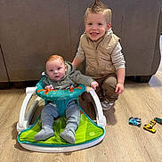 Jonah is registered to the contest to win money with this photo: baby, baby_seat, casual, child, clothing, crouching, cute, floor, hair_style, happy, home, indoor, living_room, person, playful, sibling, smiling, toddler, toy_cars, wooden_floor