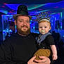 Levi is registered to the contest to win money with this photo: baby, beard, black_hoodie, blue_eyes, celebration, child, clothing, colorful_lights, facial_hair, festive, hat, headwear, holding, indoors, man, new_year, pacifier, party, smile, tattoo