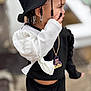 Kaioni is registered to the contest to win money with this photo: accessories, beads, black_clothing, braids, cartoon_pendant, casual, child, cute, fashion, hand_near_face, hat, long_sleeves, necklace, outdoor, portrait, profile_view, street_style, toddler, white_clothing, young_child
