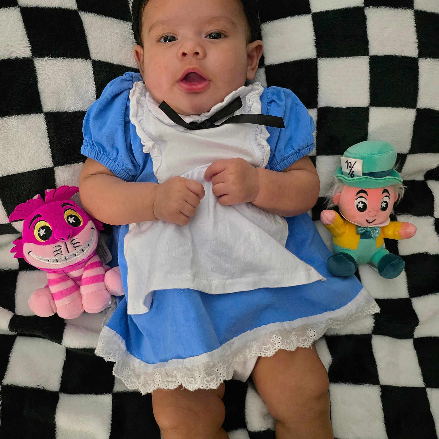 Sophia joined the competition — help win amazing prizes! baby, infant, child, portrait, lying_down, blue_dress, white_apron, black_hat, plush_toy, stuffed_animal, pink_toy, green_toy, checkerboard_blanket, smiling, face, indoor, costume, playful, photography, cute
