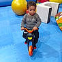 child, indoor, playground, toy_bike, blue_floor, yellow_ball, soft_play_equipment, sweater, black_pants, red_socks, curly_hair, young_child, riding, play_area, colorful, padded_floor, smiling, person, fun, leisure
