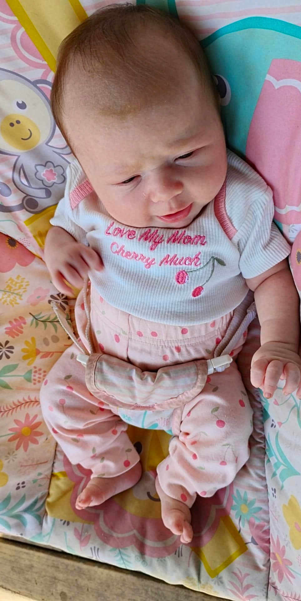 Luna is registered to the contest to win money with this photo: baby, infant, child, pink_clothing, patterned_pants, white_shirt, cherry_design, blanket, colorful, cute, indoors, sitting, footwear_free, curious_expression, soft_light, small_hands, head, face, young_child, cozy