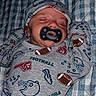 baby, sleeping, pacifier, hat, football_theme, onesie, infant, blanket, patterned_clothing, cozy, resting, cute, child, soft_lighting, indoor, portrait, newborn, face, peaceful, hands_up