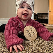 Mia joined the competition — help win amazing prizes! baby, infant, headband, bow, sweater, pink, carpet, floor, hand, milestone, wooden_sign, indoor, cute, smiling, person, child, young, portrait, crawling, home