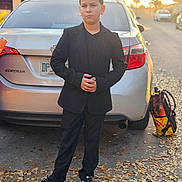 Cristian joined the competition — help win amazing prizes! child, boy, suit, black_suit, sneakers, nike, car, sedan, trunk, license_plate, backpack, street, sunset, leaves, pavement, portrait, standing, serious_expression, casual_formal_mix, urban_residential