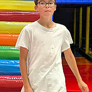 Léonid participe au concours pour gagner de l'argent avec cette photo : child, boy, glasses, white_tshirt, shorts, indoor_playground, colorful, padded_structures, walking, floor, play_area, netting, yellow, red, blue, green, orange, casual_clothing, young, person