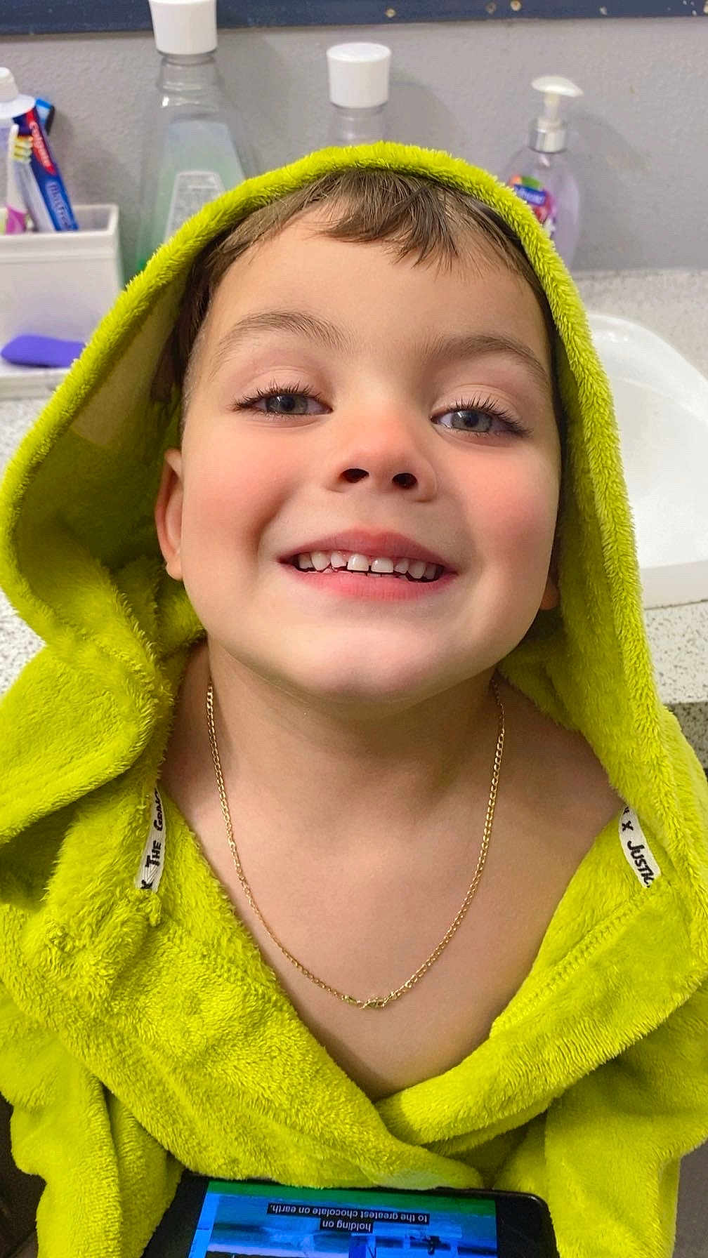 Yeison is registered to the contest to win money with this photo: child, smiling, face, hooded_towel, green_towel, gold_chain, tablet, bathroom, sink, counter, bottle, toothbrush, hygiene, young_child, portrait, happy, indoor, casual, closeup, person