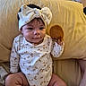 baby, child, infant, bow, headband, onesie, floral_clothing, toy, plush_toy, sitting, couch, cushion, indoor, home, portrait, smiling, face, dark_hair, cozy, photography