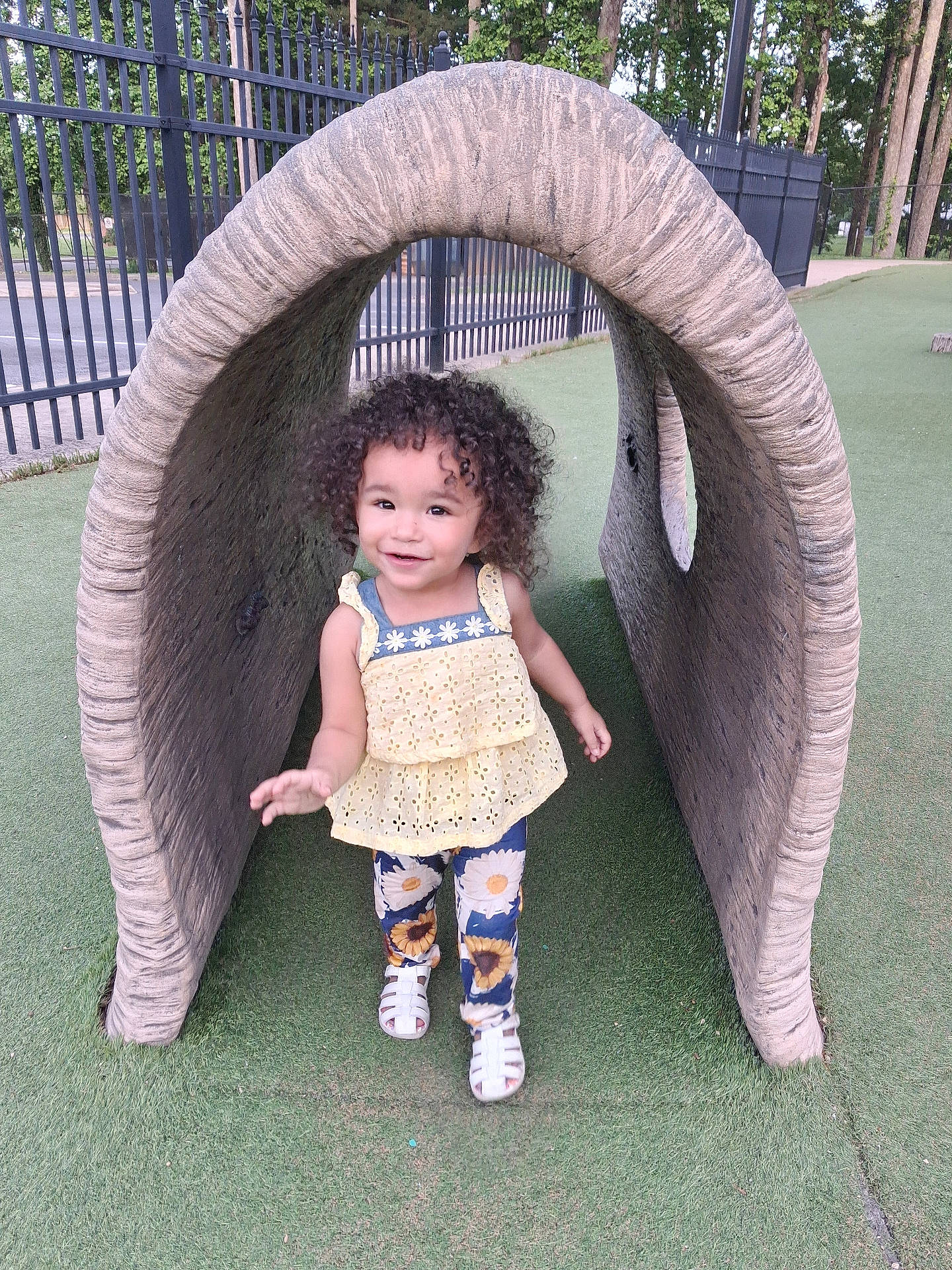 Samaria joined the competition — help win amazing prizes! artificial_turf, child, curly_hair, cute, daylight, fence, floral_pants, grass, happy, outdoor, person, play, playground, smiling, toddler, trees, tunnel, walking, white_sandals, yellow_top