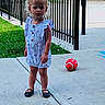 toddler, child, curly_hair, blonde_hair, dress, black_shoes, sidewalk, concrete, ball, red_ball, fence, grass, outdoor, daylight, building, brick_wall, curious_expression, person, standing, playground