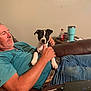 man, puppy, dog, couch, leather, blue_shirt, jeans, phone, charging_cable, table, tumbler, soda_bottle, indoor, relaxed, holding, pet, brown, black_and_white, casual, living_room