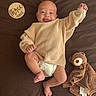 baby, infant, smile, sweater, diaper, teddy_bear, blanket, wooden_sign, brown_background, cute, happy, lying_down, raising_arm, milestone, child, face, person, cozy, soft_toy, newborn