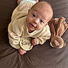 baby, infant, sweater, plush_toy, dog_paws, brown_background, wooden_sign, four_months, cute, lying_down, indoors, child, soft_toy, person, face, eyes, hands, feet, cozy, warm_colors
