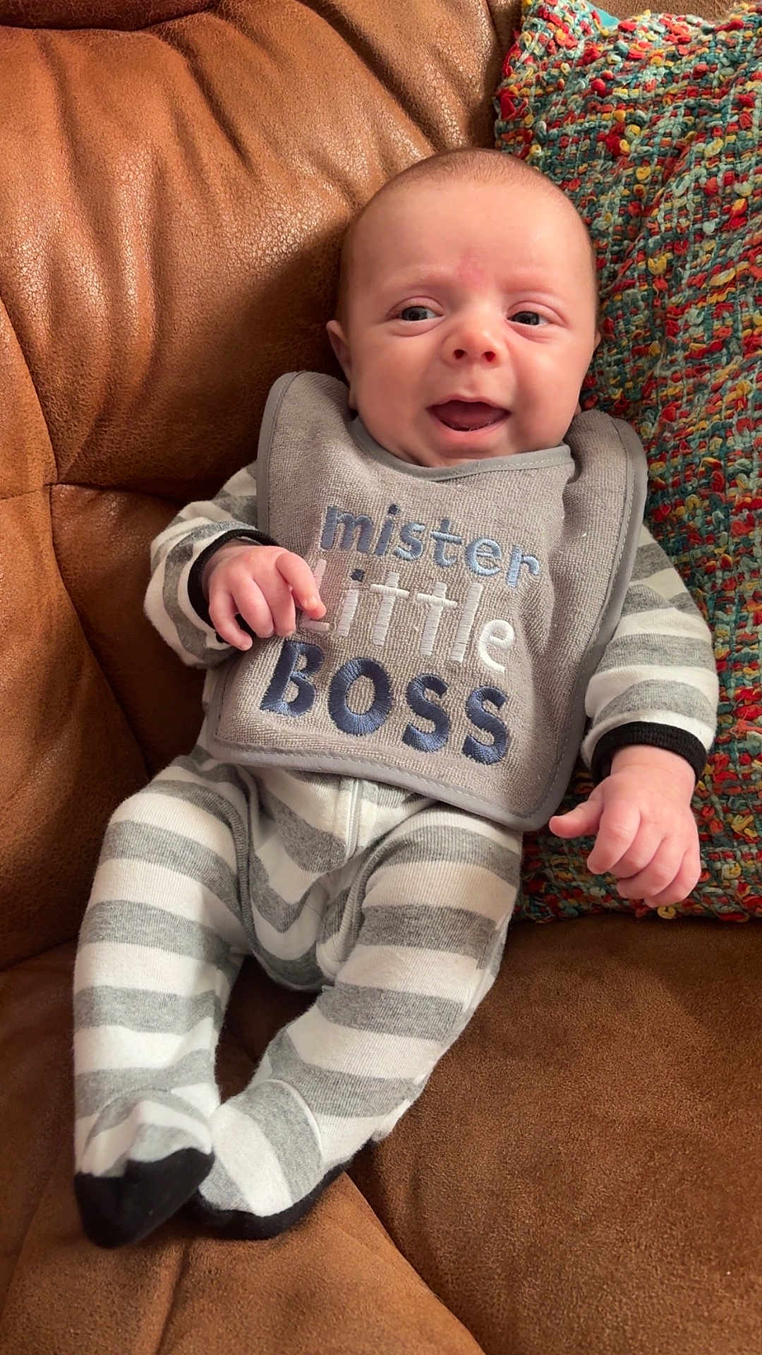 Waylon joined the competition — help win amazing prizes! baby, infant, smiling, pajamas, bib, striped_clothing, couch, brown_couch, knitted_pillow, colorful_pillow, happy, face, person, indoor, cozy, cute, small_hands, footed_pajamas, relaxing, portrait