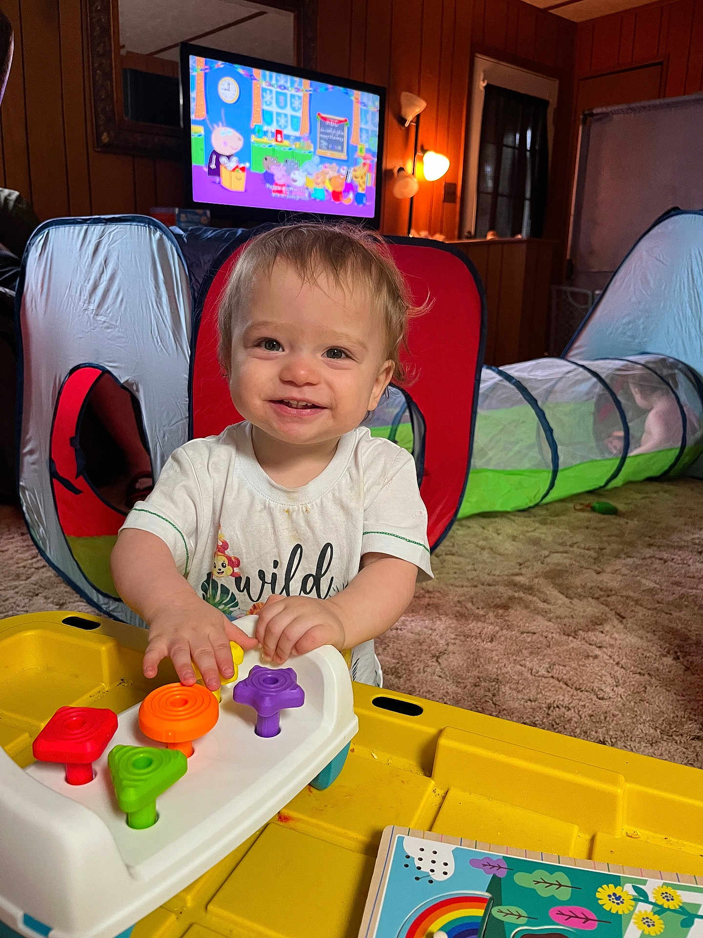 Ryland joined the competition — help win amazing prizes! toddler, child, toy, colorful_toy, yellow_table, living_room, play_tunnel, cartoon_tv, carpet, wood_paneling, smiling, happy, short_hair, indoor, furniture, lamp, book, window, playtime, person