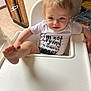Ryland joined the competition — help win amazing prizes! toddler, child, high_chair, foot, onesie, text_on_clothing, kitchen, cabinet, floor, tile, baby, person, indoor, playful, smiling, hair, seat, table, hand, expression
