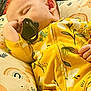 Delilah is registered to the contest to win money with this photo: baby, sleeping, pacifier, yellow_clothing, onesie, cushion, rainbow_pattern, floral_pattern, infant, peaceful, hand, ear, face, closeup, soft, comfort, rest, child, indoors, cute