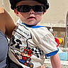 Jeremiah is registered to the contest to win money with this photo: toddler, child, sunglasses, cap, mario_kart, tshirt, kitchen, adult, hand, person, cute, indoor, fashion, casual, holding, young_child, portrait, clothing, accessory, smile