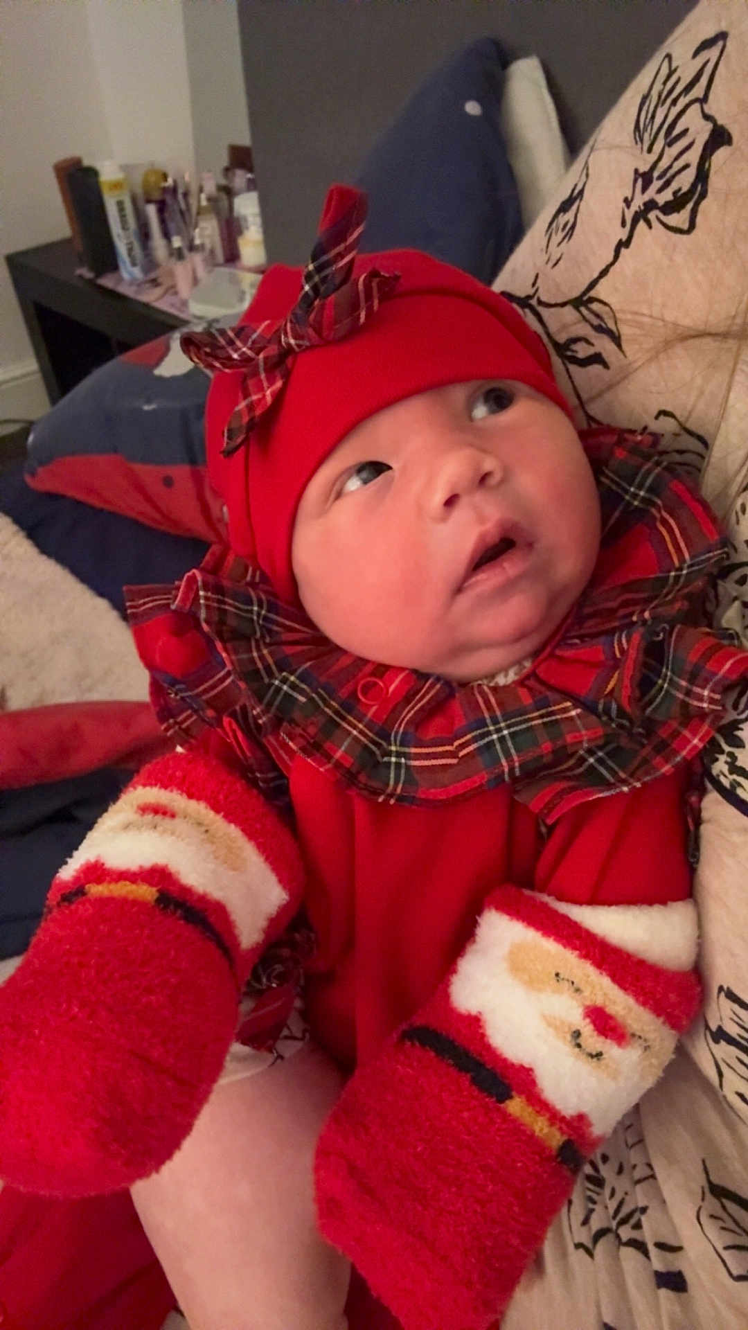 Ivy-Leigh joined the competition — help win amazing prizes! baby, infant, red_clothing, mittens, santa_mittens, plaid, hat, bow, face, cute, holiday, festive, cozy, pillow, indoors, child, person, warm_lighting, curious, expression