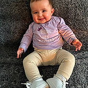Emy participe au concours pour gagner de l'argent avec cette photo : baby, child, smiling, purple_shirt, cream_pants, white_shoes, carpet, sitting, happy, face, tongue_out, indoor, cute, infant, person, young_child, plush_texture, cozy, casual_clothing, portrait