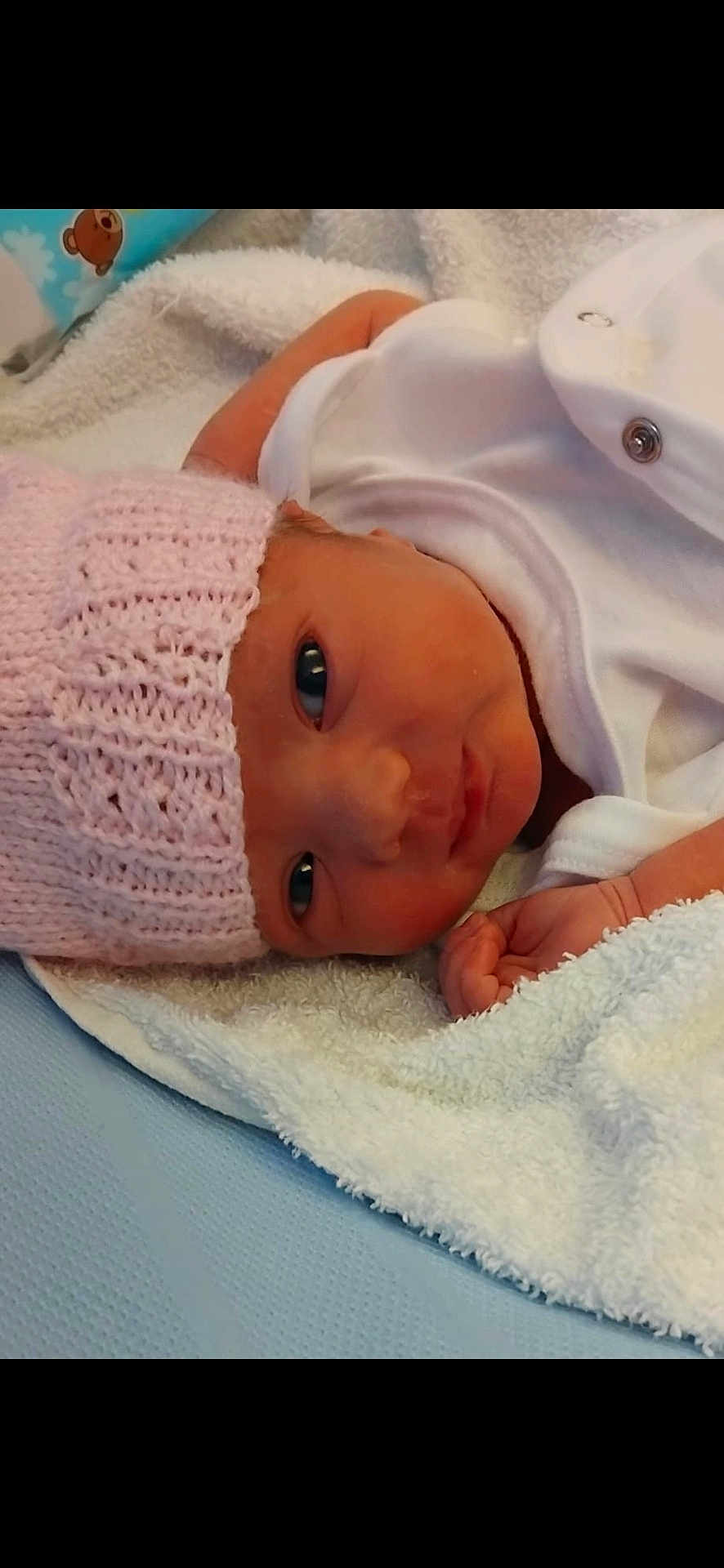 Sophia joined the competition — help win amazing prizes! newborn, baby, infant, pink_hat, knitted_hat, white_towel, soft_texture, blue_background, lying_down, face, eyes, hand, skin, cute, cozy, newborn_clothing, baby_gaze, portrait, indoors, resting