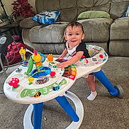 Luka is registered to the contest to win money with this photo: toddler, child, walker, toy, smile, living_room, carpet, couch, pillow, table, lamp, remote_control, colorful, playful, happy, indoor, person, footwear, baby, furniture
