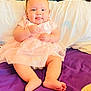 baby, infant, pink_dress, tongue_out, purple_bedspread, pillow, wooden_headboard, cute, smiling, sitting, indoors, child, soft_lighting, barefoot, chubby_cheeks, happy, portrait, tulle_skirt, relaxed, cozy
