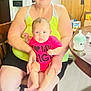woman, baby, glasses, chair, kitchen, refrigerator, baby_bottle, wood_paneling, table, onesie, sitting, indoor, person, child, smiling, home, furniture, magnet, artwork, casual_clothing