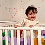baby, crib, child, smiling, curly_hair, foam_puzzle, heart_stickers, white_crib, indoor, happy, infant, play_area, colorful, puzzle_pieces, wall_decor, hand_grip, standing, cute, room, young_child