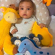 Zohan is registered to the contest to win money with this photo: baby, child, plush_toy, giraffe, dinosaur, stuffed_animal, cushion, bed, pajamas, infant, cute, indoor, soft_toy, person, face, feet, star_pattern, yellow, blue, orange