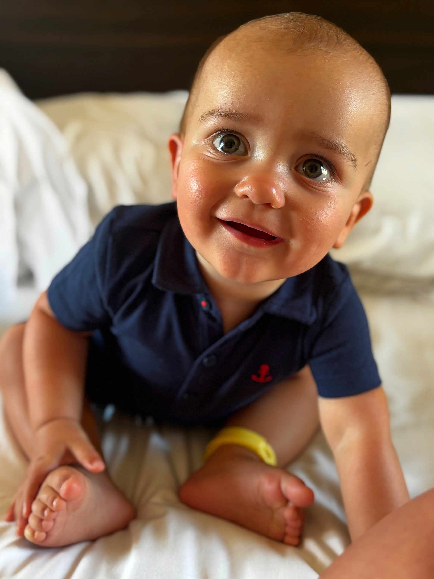 Freddie joined the competition — help win amazing prizes! baby, child, smiling, sitting, navy_blue_shirt, anchor, bed, white_bedspread, feet, hands, happy, cute, infant, portrait, indoors, closeup, skin, expression, person, young_child