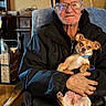 black_jacket, blue_collar, casual_clothing, chair, companionship, cozy, dog, elderly_man, glasses, hand, indoor, pet, portrait, relaxed, rustic_furniture, seated, small_dog, smile, warm_lighting, wooden_furniture