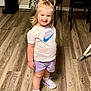 casual_clothing, chair, child, colorful_sneakers, cute, flooring, furniture, happy, home_interior, indoor, person, pony_tail, purple_shorts, smiling, standing, table, toddler, white_shirt, wooden_floor, young_child