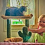 Sofia is registered to the contest to win money with this photo: toddler, child, cat, cat_tree, window, curtain, green_dress, indoor, furniture, plant_shaped_cat_tree, light, smile, face, person, pet, animal, home, playful, cute, cozy