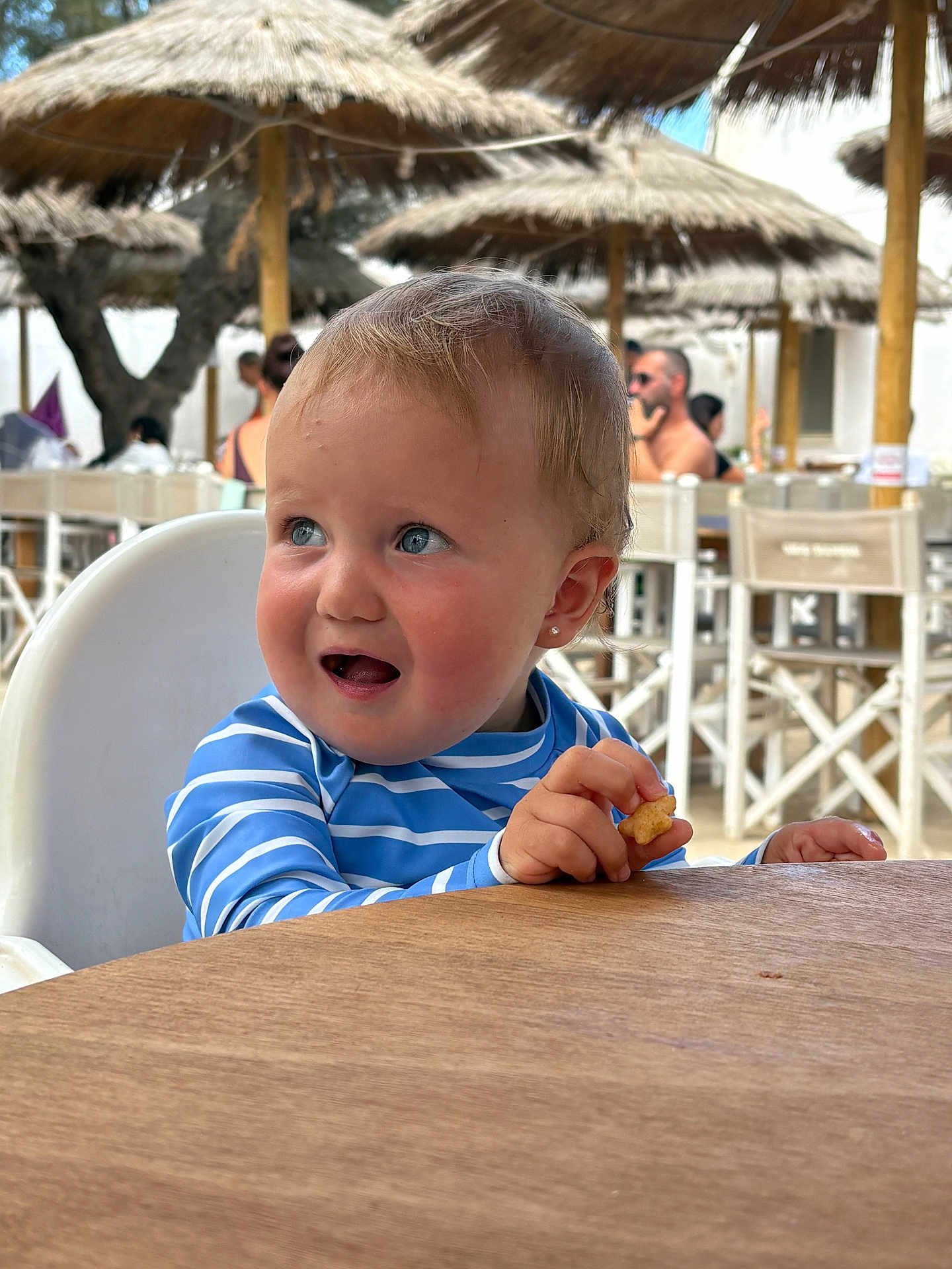 Sofia joined the competition — help win amazing prizes! toddler, child, high_chair, blue_eyes, snack, striped_shirt, outdoor, restaurant, beach_umbrella, summer, daylight, people, table, wooden_table, casual, expression, eating, cute, baby, portrait
