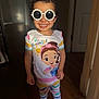 child, toddler, smiling, sunglasses, curly_hair, pajamas, barefoot, indoor, colorful_clothing, cartoon_print, happy, portrait, standing, floor, wooden_floor, door, light, shadow, cute, playful