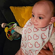 Juliette a rejoint le concours — aidez-le/la à gagner de superbes lots ! baby, bib, child, closeup, clothing, couch, cute, eyes, face, hand, heart_pattern, holding, indoor, infant, person, pink_outfit, seated, soft_lighting, toy, yellow_pillow
