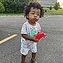 toddler, child, watermelon, curly_hair, outdoor, parking_lot, asphalt, shorts, tshirt, crocs, food, summer, greenery, building, sunset, casual_clothing, cute, messy_face, holding, standing