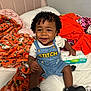 Tylin is registered to the contest to win money with this photo: toddler, child, smiling, curly_hair, denim_overall, sitting, bed, blanket, orange_blanket, pink_blanket, pillow, pack_of_wipes, pacifier, black_shoes, indoor, happy, person, clothing, baby, hand