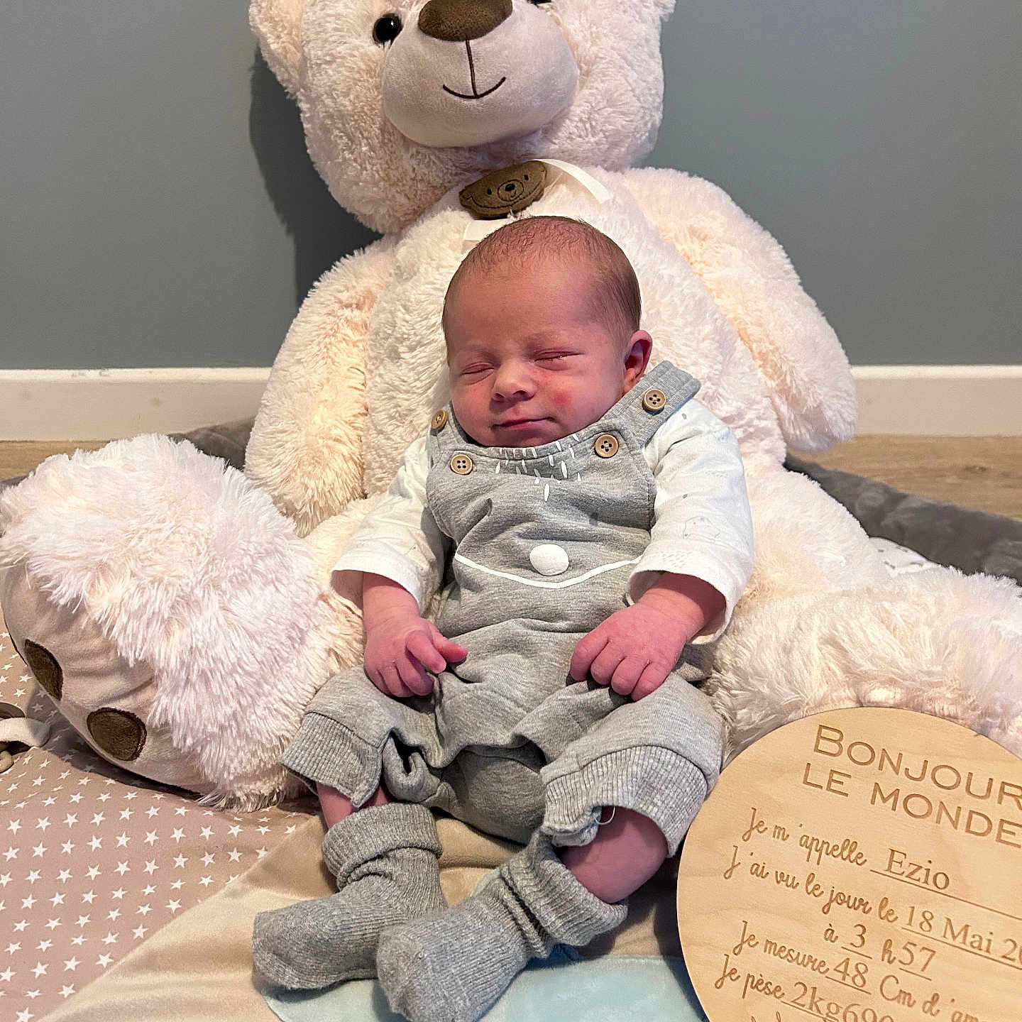 Ezio participe au concours pour gagner de l'argent avec cette photo : baby, blanket, child, cute, floor, grey_overalls, homedecor, indoor, infant, newborn, person, plush_toy, portrait, sleeping, smiling, socks, soft_toy, star_pattern, teddy_bear, wooden_plaque